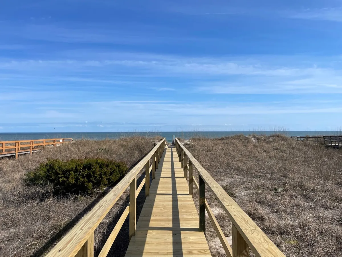 A serene view along a wooden walkway leading through dune grass toward the ocean, with gentle waves visible in the background. The walkway invites guests directly onto the sandy beach, evoking the anticipation of a beach day.