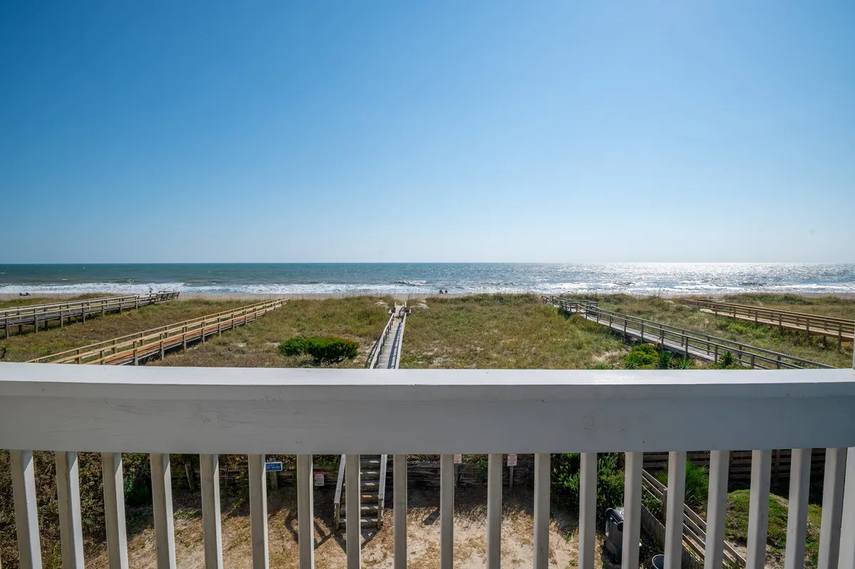 A wide, panoramic view of the ocean framed by sandy dunes and boardwalks, with the beach stretching into the distance. The scene captures the golden light of either sunrise or sunset, evoking peacefulness and opportunities for romantic walks or beachcombing.