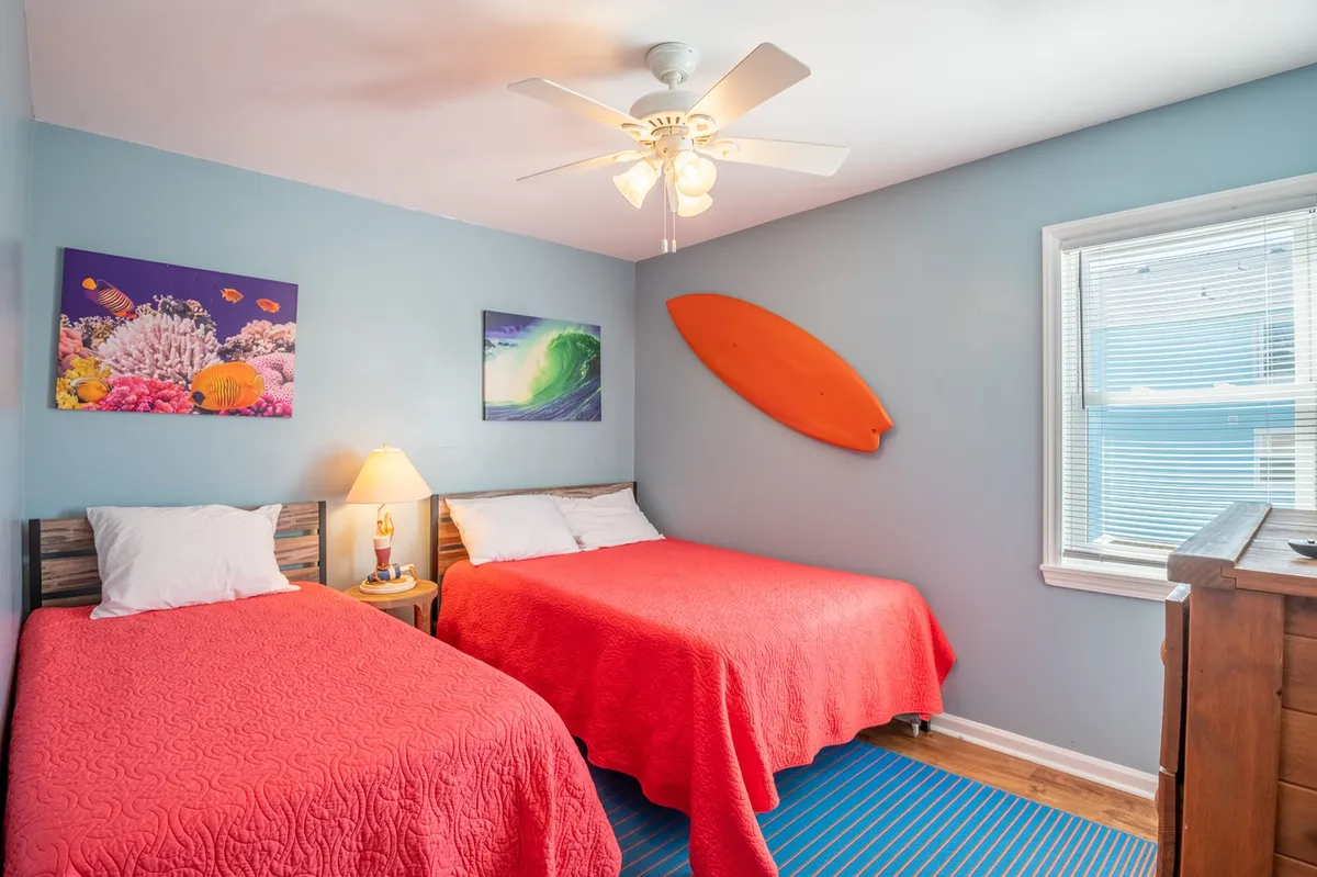 This image features a guest bedroom with two beds adorned in coral-colored bedspreads, colorful marine-themed wall art, a surfboard decoration, and a wooden dresser. Sunlight streams through the window, adding warmth to the playful, family-friendly décor.