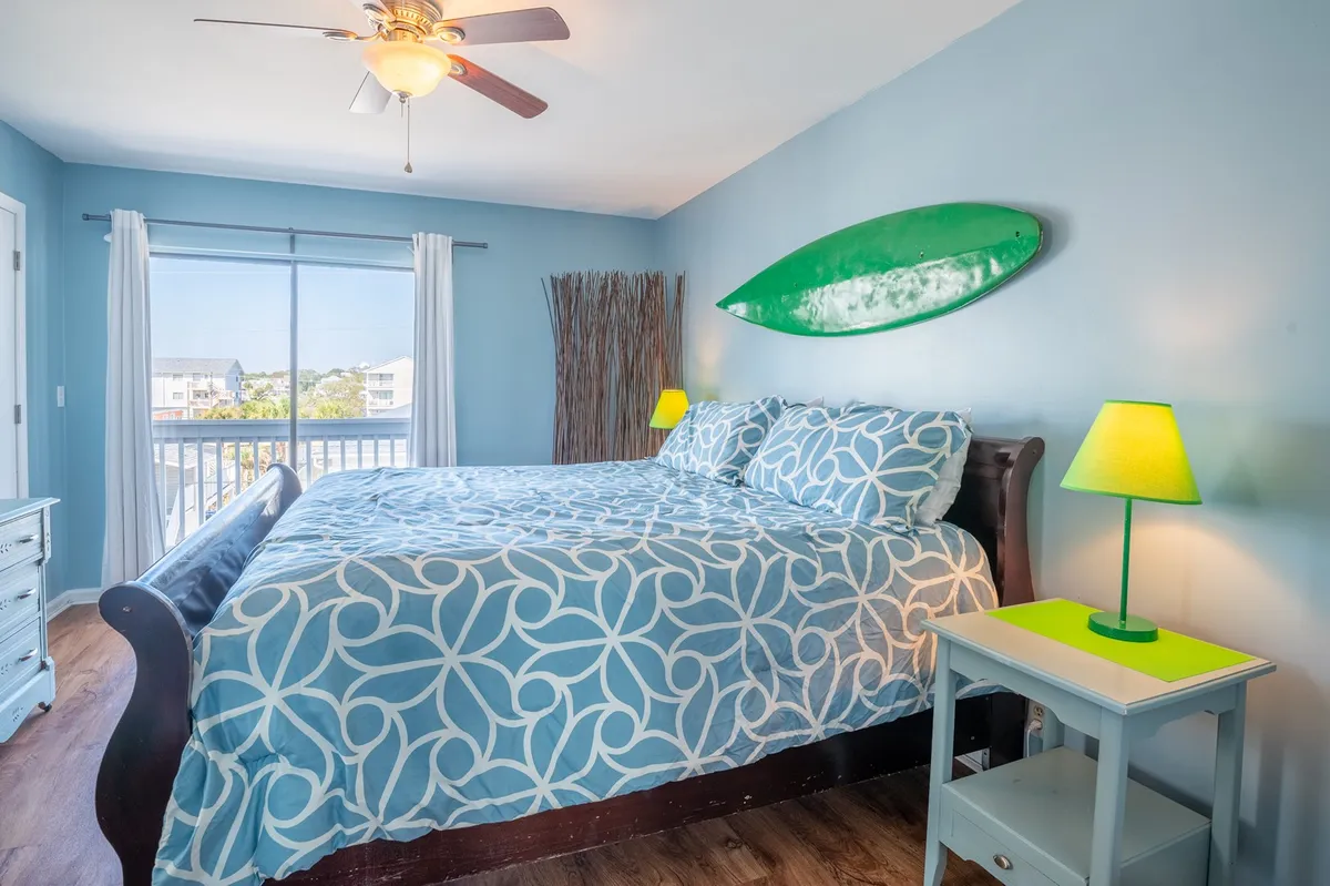 This image displays a bedroom with a bed featuring blue patterned bedding, a green surfboard mounted on the wall, and a yellow bedside lamp. Natural light streams in, creating a bright and inviting atmosphere that feels both playful and relaxing.