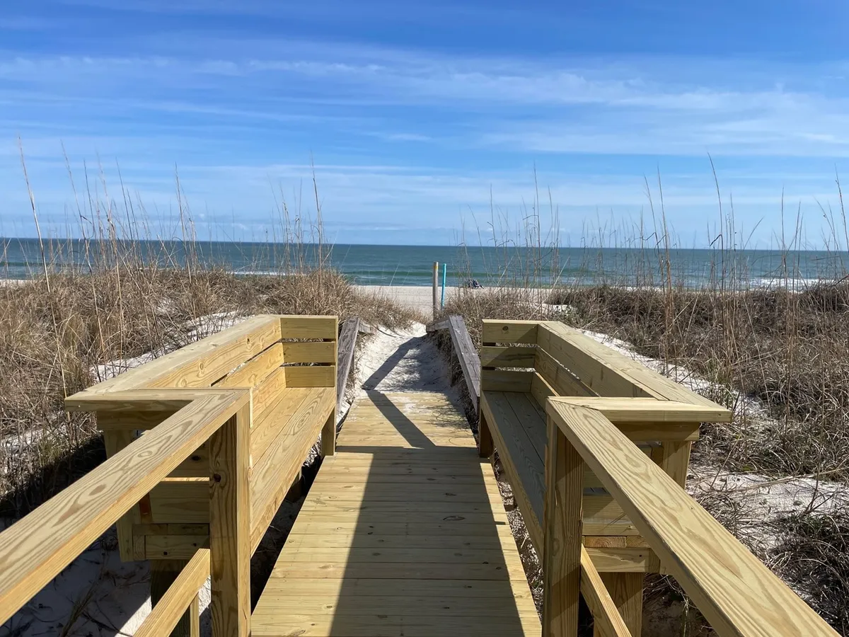 A wooden boardwalk stretches through beach grass toward the ocean, offering a relaxed and inviting pathway for guests. The open view of the beach and water beyond highlights the proximity to the shore and suggests easy access for sunset strolls or moonlit walks described in the section.
