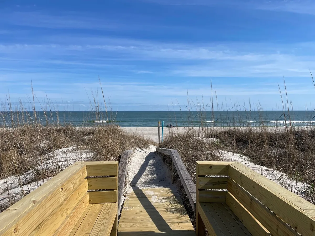 A tranquil exterior shot featuring a private wooden walkway leading through the dunes directly to the sandy beach and ocean beyond. The path is lined with beach grass and feels secluded, emphasizing the privacy and exclusivity of the community beach access.