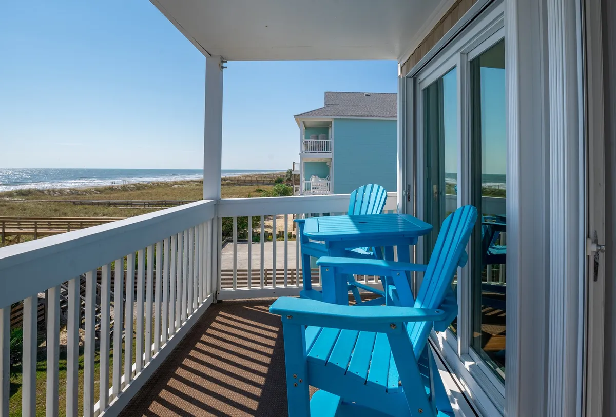 An exterior shot of the balcony with blue Adirondack chairs overlooking the ocean. The railings frame a sweeping coastal view, evoking the feeling of fresh air and tranquility. The scene suggests sipping coffee or watching sunsets while enjoying the sound and breeze of the waves.