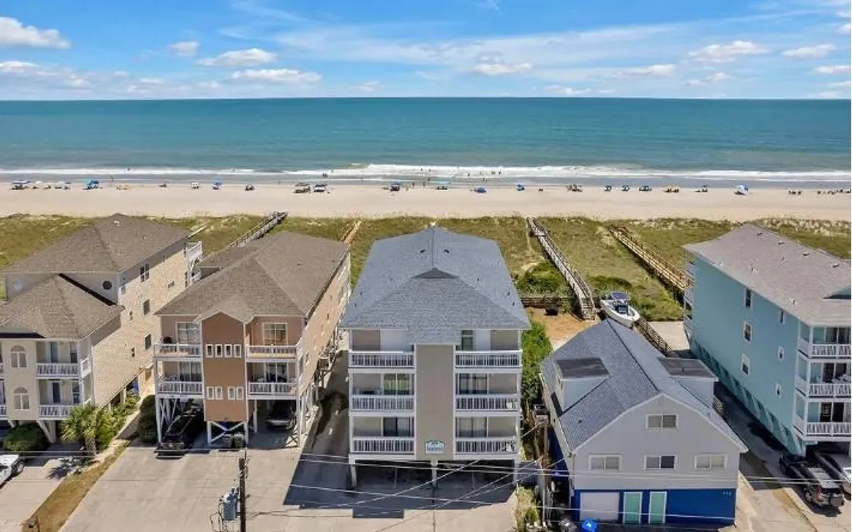 A sweeping exterior view features the oceanfront with gentle waves, sandy shores, and neighboring homes. The angle highlights the proximity to the water, communal spaces, and opportunities for a variety of family beach activities, from morning walks to evening gatherings.