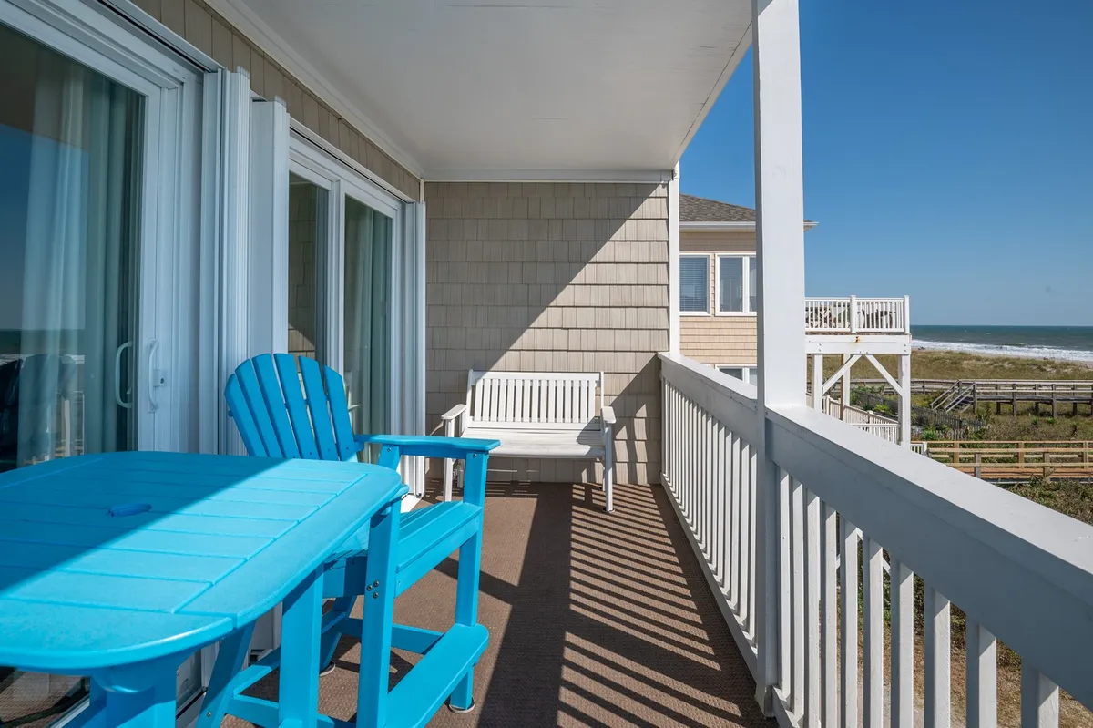 A serene exterior scene featuring blue Adirondack chairs and a white bench set on a balcony or patio overlooking the ocean. The sandy beach and rolling waves are visible beyond, inviting guests to relax and savor the view. The setting is perfect for sipping morning coffee or unwinding at sunset, encapsulating privacy and romance.