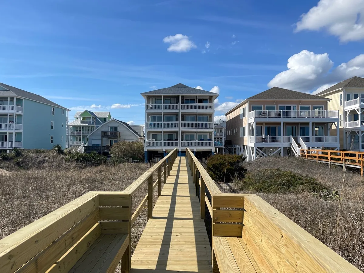 A charming wooden walkway leads from the home through soft sand dunes toward a beautiful expanse of beach, framed by inviting beach houses and a vibrant blue sky overhead. The scene captures the anticipation and excitement of heading out to the beach for a day of family fun.