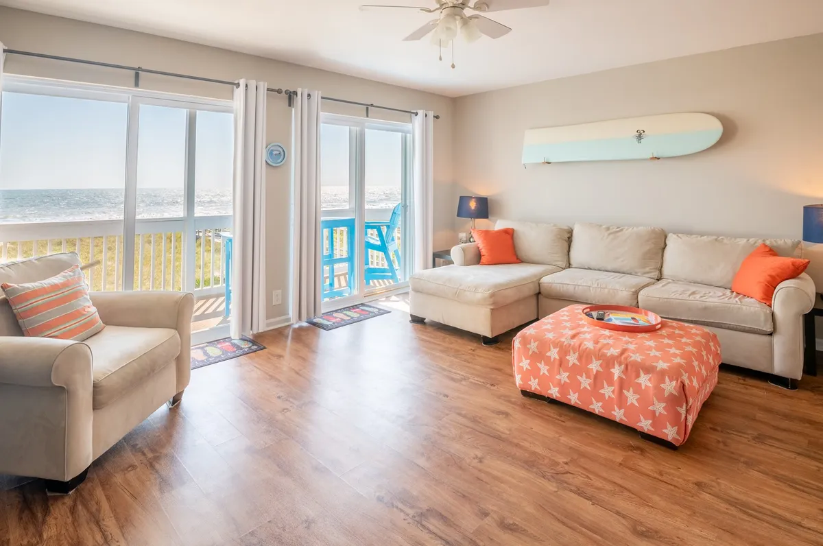 A bright, inviting living room with a comfortable sectional sofa and a colorful ottoman, all oriented toward expansive windows framing breathtaking ocean views. The mood is cheerful and laid-back, perfect for relaxing, socializing, or simply savoring the sunrise over the Atlantic.