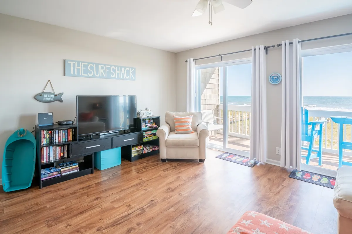 A casual, beach-themed living area with a television, comfortable armchair, large windows, and relaxed decor. The room feels bright and welcoming, perfect for relaxing, working remotely, or enjoying movie nights.