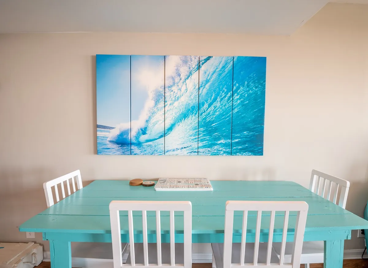 A bright, airy dining area features a large wave artwork on the wall, a turquoise dining table, and white chairs, exuding a casual coastal vibe. Windows let in abundant natural light, tying the interior scene to the oceanfront setting mentioned in the section.