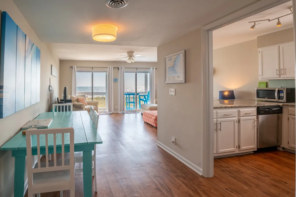 An open-plan interior scene featuring a dining table, living area with seating, kitchen entrance, and large windows offering an expansive ocean view. The natural light floods the space, enhancing the modern yet cozy aesthetic. The layout encourages communal activities and relaxation while maintaining a strong visual connection to the coast.